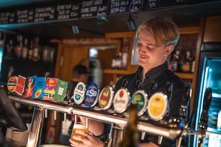 A beer bar counter with several taps and a staff member pouring beer into a glass. A warm atmosphere with dim, moody lighting.