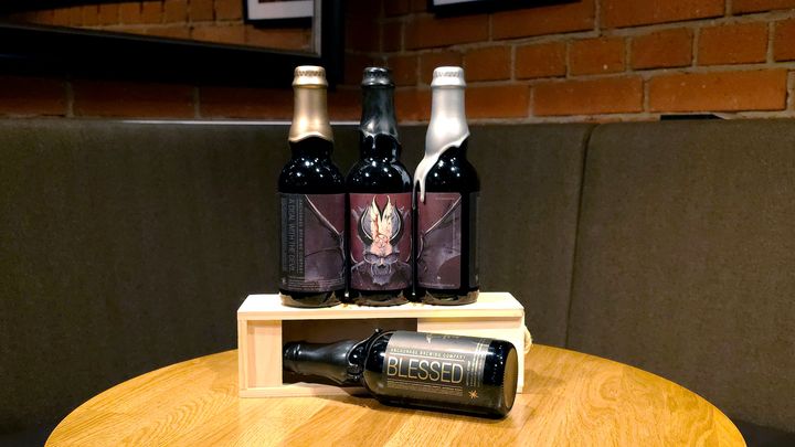 Alt text: Bottles of Anchorage Brewing Company beers, including A Deal With the Devil – Penta Oaked, Batch 1–3, standing upright on a wooden stand, and Blessed Imperial Stout lying horizontally on a table.