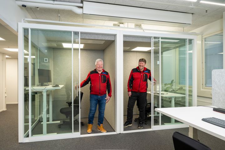Ilkka Okkonen ja Pasi Jakonen lasiseinäisten työhuoneiden ovella. Toimistomiljöö. Molemmilla on punaiset pusakat ja leveä hymy.