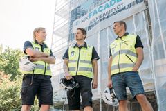 Kolme henkilöä julkisivuremontissa olevan rakennuksen edustalla, rakennustelineiden edessä. Heillä on turvaliivit yllä ja kypärät kädessä.