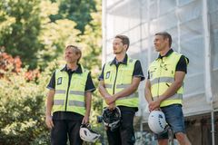 Kolme henkilöä julkisivuremontissa olevan rakennuksen edustalla, rakennustelineiden edessä. Heillä on turvaliivit yllä ja kypärät kädessä.