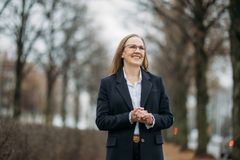 Eeva Riitta-Tuomisen puolivartalokuva. Ilme on hymyilevä, katse ylävistoon ja kädet ovat edessä yhteenliitettyinä. Hänellä on yllään tumma pitkä takki, joka on auki. Taustalla syksyinen puistokuja, lehdet ovat jo pudonneet puista.