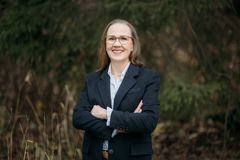 Puolivartalokuva Eeva-Riitta Tuomisesta. Katse on kameraan, kädet ovat ristissä rinnalla, ilme hymyilevä. Hänellä on yllään tumma pitkä takki, joka on auki. Taustalla puistomaisema.