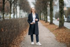 Kokovartalokuva Eeva-Riitta Tuomisesta sykyisellä puiston hiekkatiellä, ilme on hymyilevä ja katse kameraan. Asento on oikealle jalalle nojaava, oikea käsi on taskussa. Hänellä on yllään tumma pitkä takki, joka on auki.  Puut ovat jo lehdettömiä.