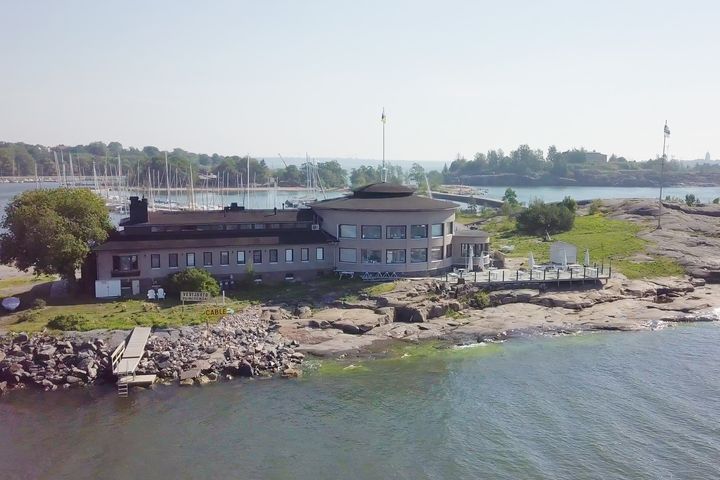 Rakennus meren rannalla, jossa on pyöreä ravintolasali. Näkymä on kesäinen ja taustalla näkyy purjeveneitä ja saaristoa.