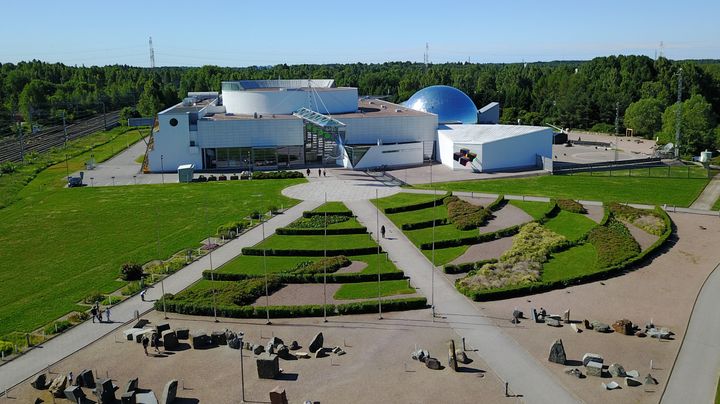 Ilmakuva tiedekeskuksesta, jonka ympärillä on vehreää puistoaluetta ja kasvien muotoon istutettuja pensaita.