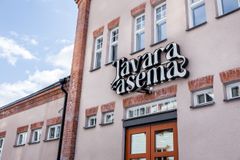 Tavara-aseman maitokahvin värinen julkisivu kuvattuna sinistä taivasta vasten. Talon oven päälle on ripustettu Tavara-aseman logon muotoinen valokyltti.