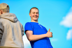 En ung kille i blå t-skjorta visar tummen upp mot blå himmel i bakgrunden. En annan kille står vänd med ryggen åt.