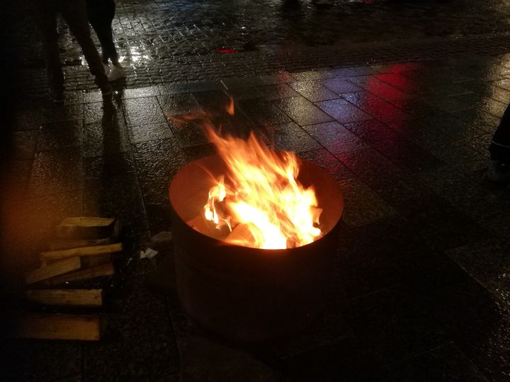 Kuvassa palaa asunnottomien yön tuli metallityynyrissä sateisessa pimeässä katumaisemassa.