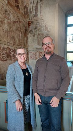 En kvinna och en man står i Esbo domkyrka bredvid varandra och bakom skymtar kyrkans kalkmålningar i brunt.