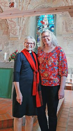 Två kvinnor står bredvid varandra i Esbo domkyrka med en skymt av altarglasmålningen i bakgrunden.