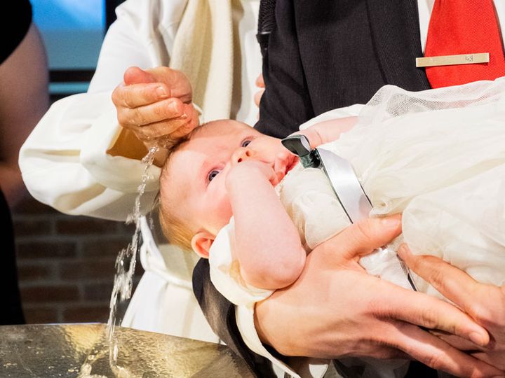 Ett spädbarn hålls i dopklänning intill dopfunten och döps av en präst.