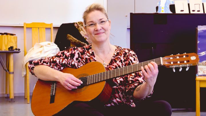 En kvinna med blont hår och glasögon sitter på golvet med en gitarr i famnen.