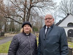 En kvinna och en man i mörka kläder står på en kyrkogård med träd och gräsmatta i bakgrunden.