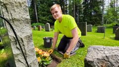 En ung man med ljust hår iklädd en gul skjorta och svarta byxor sitter på huk intill en gravvård och sköter blomplantering.