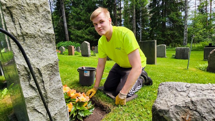 En ung man med ljust hår och klädd i gul skjorta och svarta byxor sitter på huk intill en gravvård och sköter blomplantering.