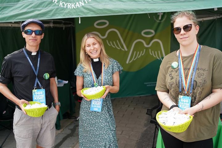 En man och två kvinnor står framför ett grönt tält med skålar i händerna, för att dela ut något till besökare.