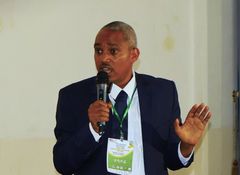 En mörk man står i kavaj och talar i en mikrofon.