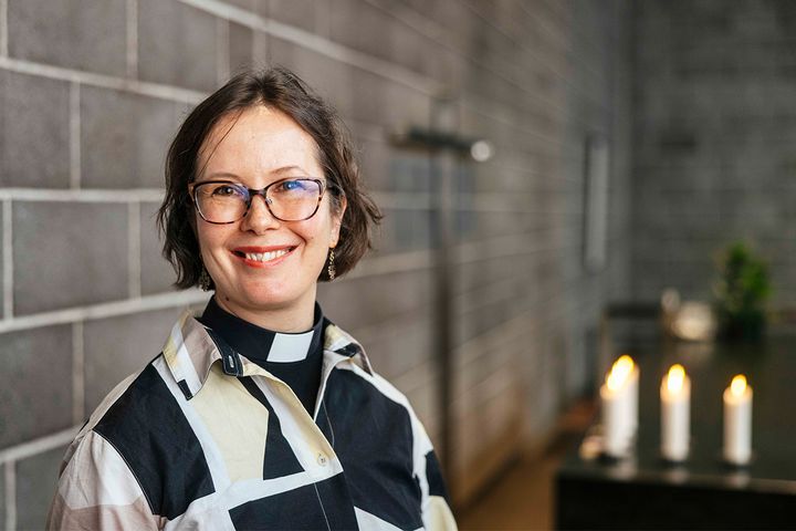 En leende kvinna med pageklippt ljusbrunt hår och glasögon fotograferad i ett kyrkorum med grå tegelvägg och tända ljus.