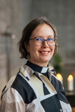 En leende kvinna i pageklippt ljusbrunt hår och glasögon står intill en grå tegelvägg och ett par tända ljus.
