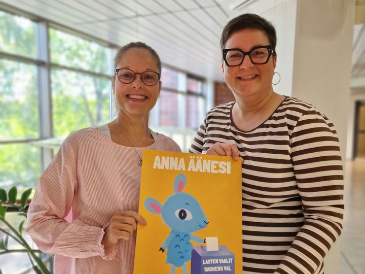 Två kvinnor håller i en affisch med uppmaningen: ge din röst och en illustration av ett lamm som lägger en valsedel i en valurna.