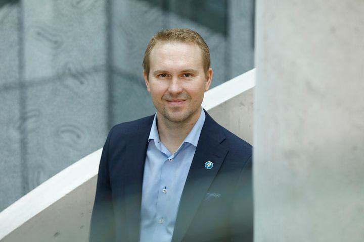 Mies hymyilee pukeutuneena pukuun ja seisoo sisätiloissa.