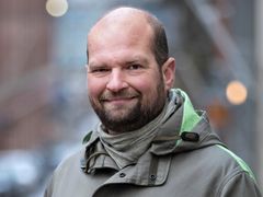 Kuvassa Sampo Ruoppila hymyilee ja katsoo kameraan. Hänellä on takki ja taustalla näkyy kaupunkinäkymää.