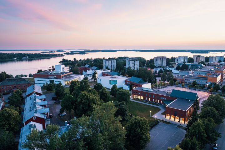 Ilmakuva Vaasan yliopiston kampuksesta meren rannalla, rakennuksia ja puita auringonlaskun aikaan.