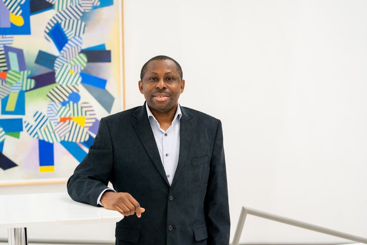 Pius Ewoh seisoo valkoisen taustan edessä, pukeutuneena tummapukuiseen takkiin, ja taustalla on värikäs, geometrinen maalaus seinällä.