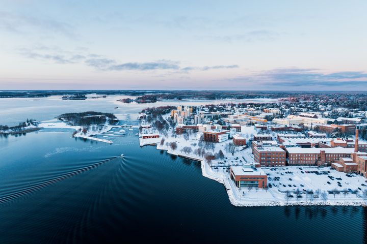 Ilmakuva talvisesta kampuksesta veden äärellä, lumipeitteisiä rakennuksia ja veneen jättämiä jälkiä vedessä.