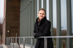 Helka Kalliomäki seisoo rakennuksen edessä, nojaa kaiteeseen ja katsoo kameraan.