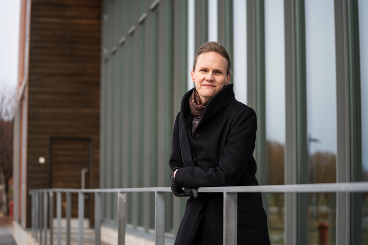 Helka Kalliomäki seisoo rakennuksen edessä, nojaa kaiteeseen ja katsoo kameraan.
