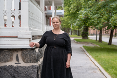 Salla Maijala seisoo ulkona puna-valkoisen vanhan puutalon edessä, ympärillä puita.