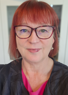 Marita Ahola, punatukkainen nainen, jolla on silmälasit ja hymyilevä ilme, pukeutuneena mustaan kuviolliseen takkiin ja pinkkiin paitaan.