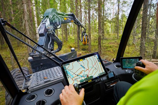 Näkymä metsäkoneen ohjaamosta, josta käsin ihminen ohjaa koneen puomia. Näytöillä näkyvät kartta ja kamera.