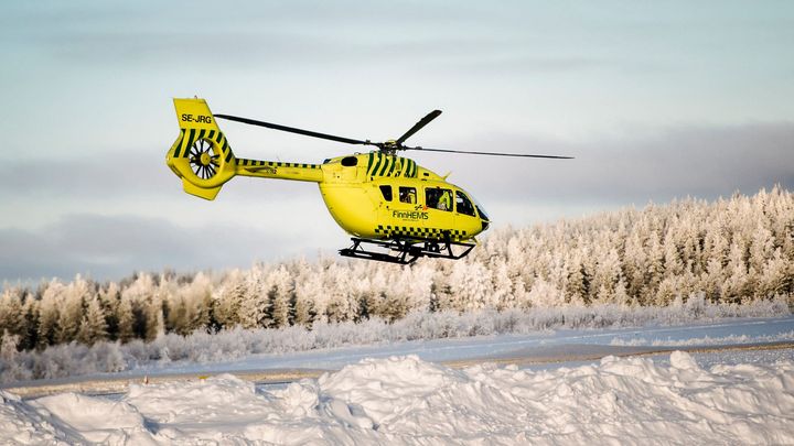 Keltainen lääkärihelikopteri lentää lumisen metsämaiseman yllä.