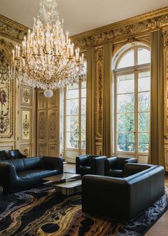 Alt-teksti: Elegantti huone, jossa on kultaisia yksityiskohtia, suuri kristallikattokruunu ja mustia nahkasohvia. Huoneessa on suuret kaari-ikkunat.