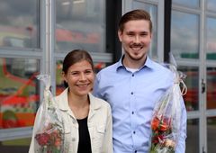 Hymyilevät nuori nainen ja mies seisovat kukkakimppujen kanssa pelastuslaitoksen kalustohallin lasiovien edessä.