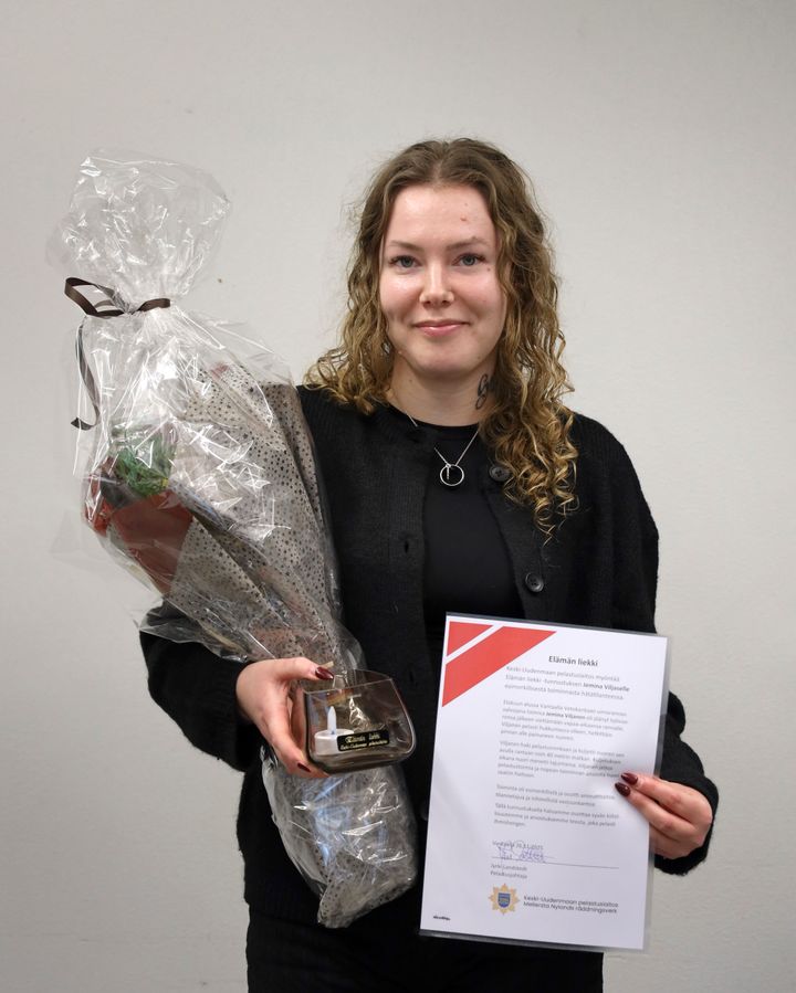 Hymyilevä nainen pitää käsissään suurta kukkakimppua ja kunniakirjaa.