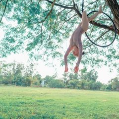 A person in a leotard performs an aerial hoop pose, hanging upside down from a tree branch in a park.