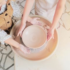 Two people are shaping a clay bowl on a pottery wheel.