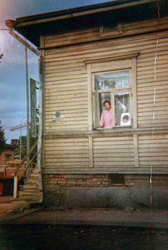 Vanha valokuva puutalosta 1970-luvulta, jossa kaksi ihmistä katsoo ulos ikkunasta. Talo on väriltään