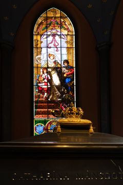 Värikäs lasimaalaus kirkossa, jossa on kuvattuna useita ihmishahmoja. Edustalla on koristeltu arkku.