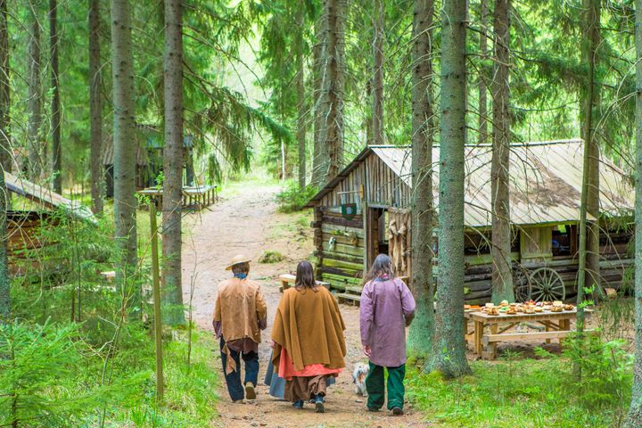 Kolme henkilöä kävelee metsätiellä selkä kameraan päin kohti puisia rakennuksia.