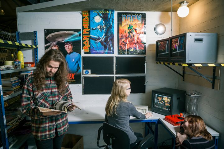 Kolme henkilöä retrohenkisessä huoneessa, jossa on vanhoja tietokoneita ja seinillä värikkäitä julisteita.