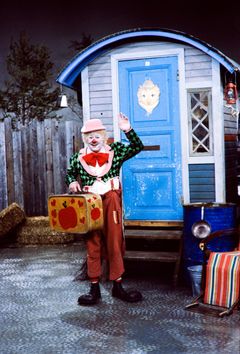 Pelle Hermanni seisoo sinisen asuntovaunun edessä, kädessään keltainen matkalaukku, toinen käsi kohotettuna tervehdykseen.
