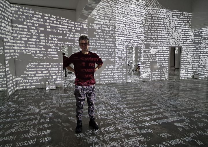 Mies seisoo hämärässä huonetilassa, jonka seinille on heijastettu tekstiä.