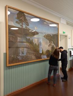 Kaksi henkilöä tutkii seinällä olevaa isoa maisemamaalausta.