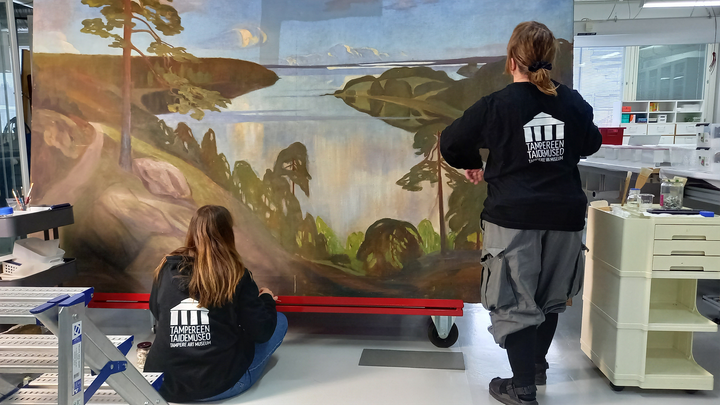 Kaksi mustapuseroista naista tutkii isoa maisemamaalausta.