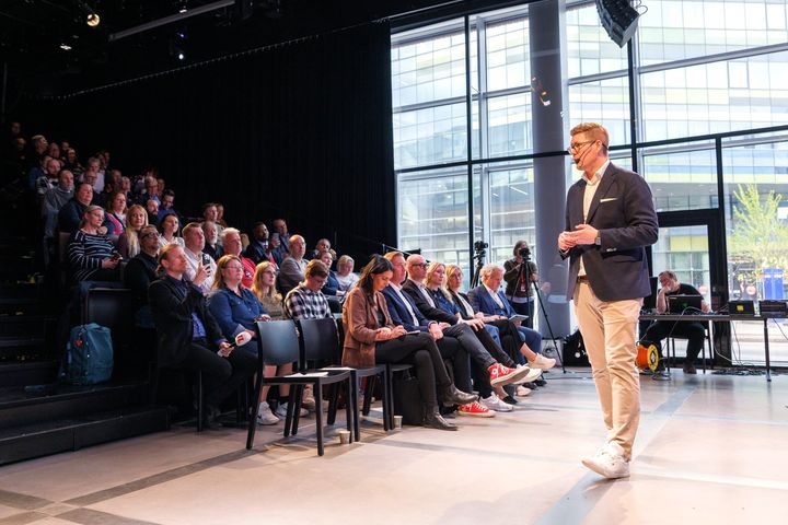 SDP:n puheenjohtaja Antti Lindtman puhumassa yleisölle Oodin tapahtumassa. Kuvan yhteydessä korostetaan Lindtmanin viestiä siitä, että tasa-arvo, koulutus ja hyvinvointipalvelut ovat Suomen menestyksen ja kilpailukyvyn perusta.